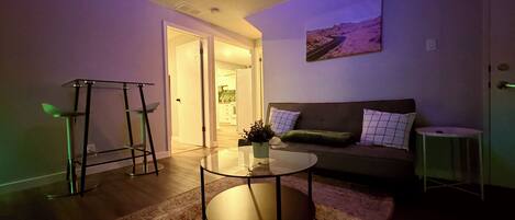 A living room with a glass coffee table, a sofa with cushions, a painting on the wall, and a door leading to another room.