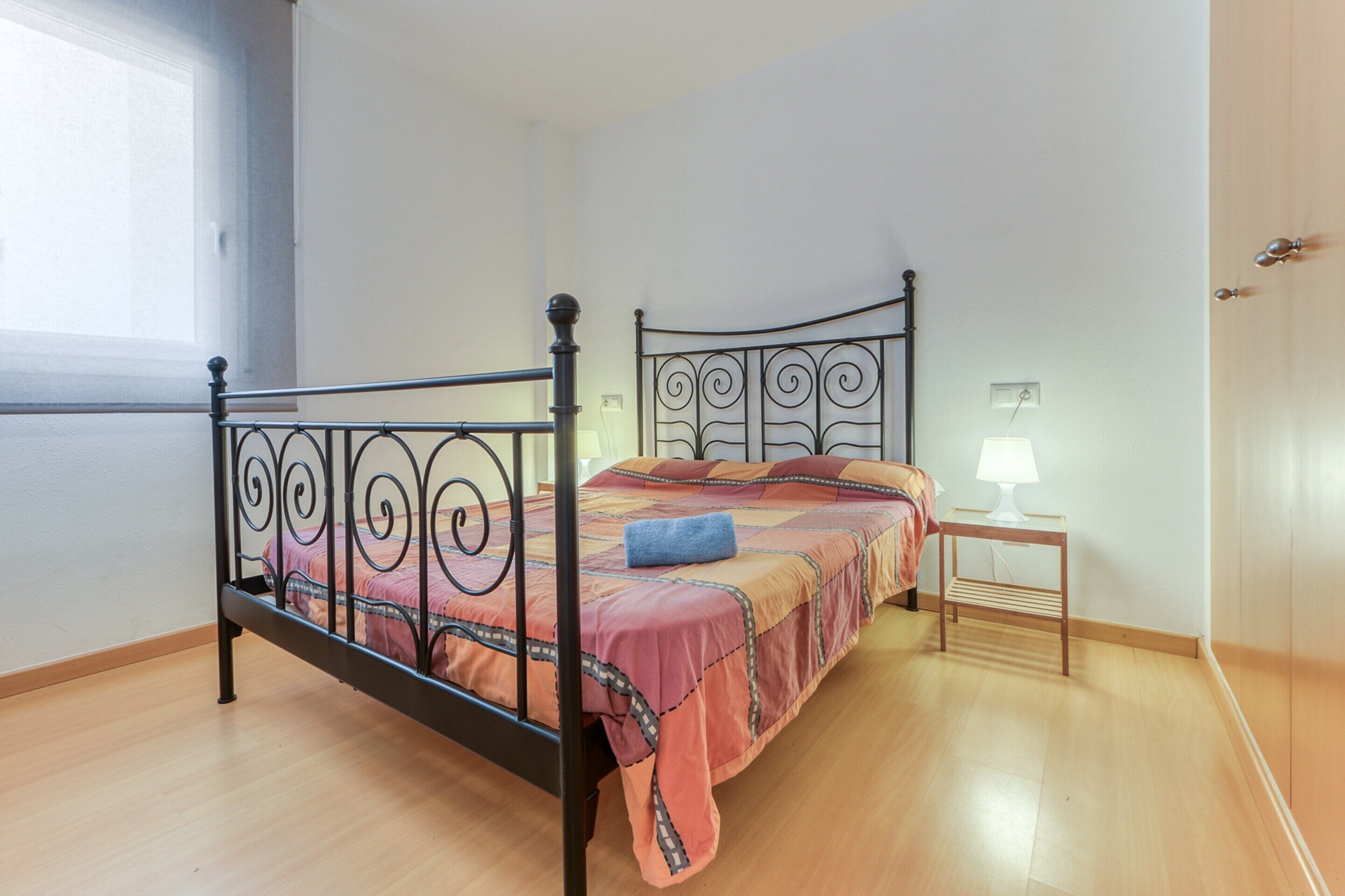 Un dormitorio con estructura de cama metálica, mesita de noche de madera con lámpara, y ventana que proporciona luz natural.