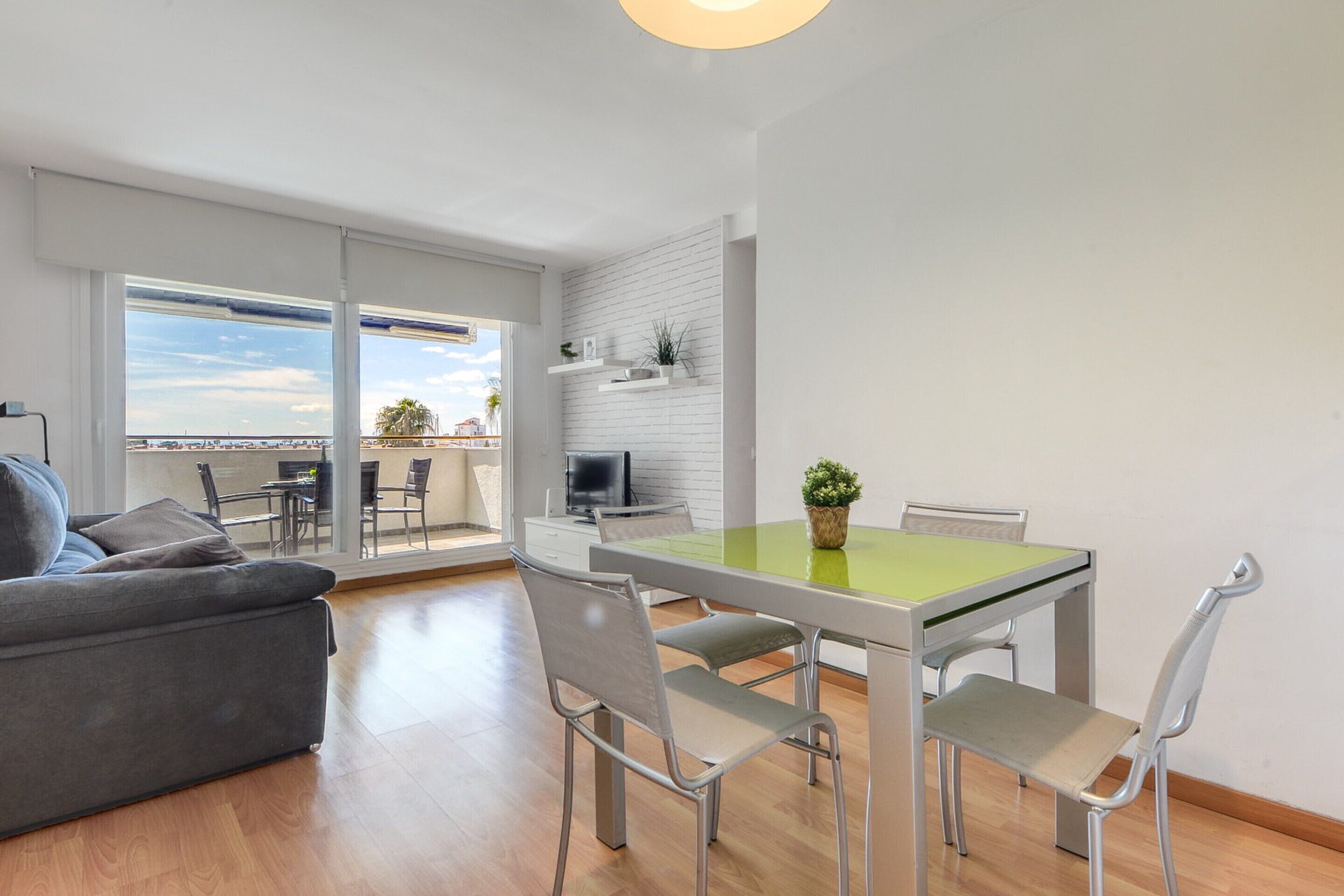 A modern living room with a grey sofa, a dining table with chairs, a TV, and a balcony with a view of the beach.
