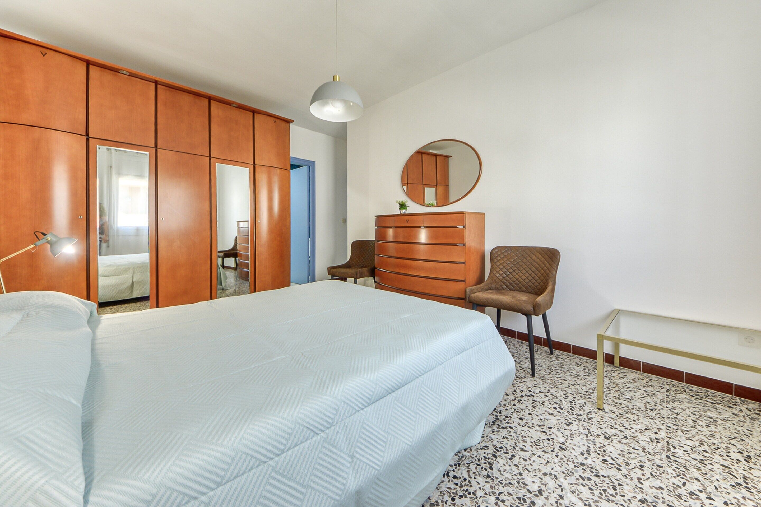 Ein Schlafzimmer mit Bett, Kommode, Stuhl, Spiegel und Pendelleuchte.