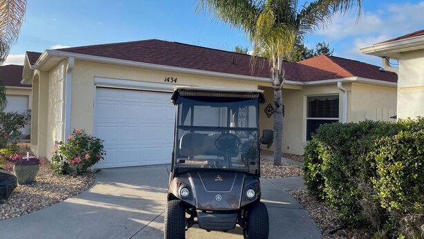 A golf cart parked in front of a house with a garage, a palm tree, and a hedge.