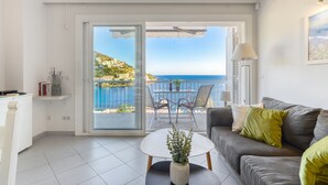 Una sala de estar moderna con un sofá gris, una mesita redonda con una planta y vista al mar a través de un ventanal corredero.