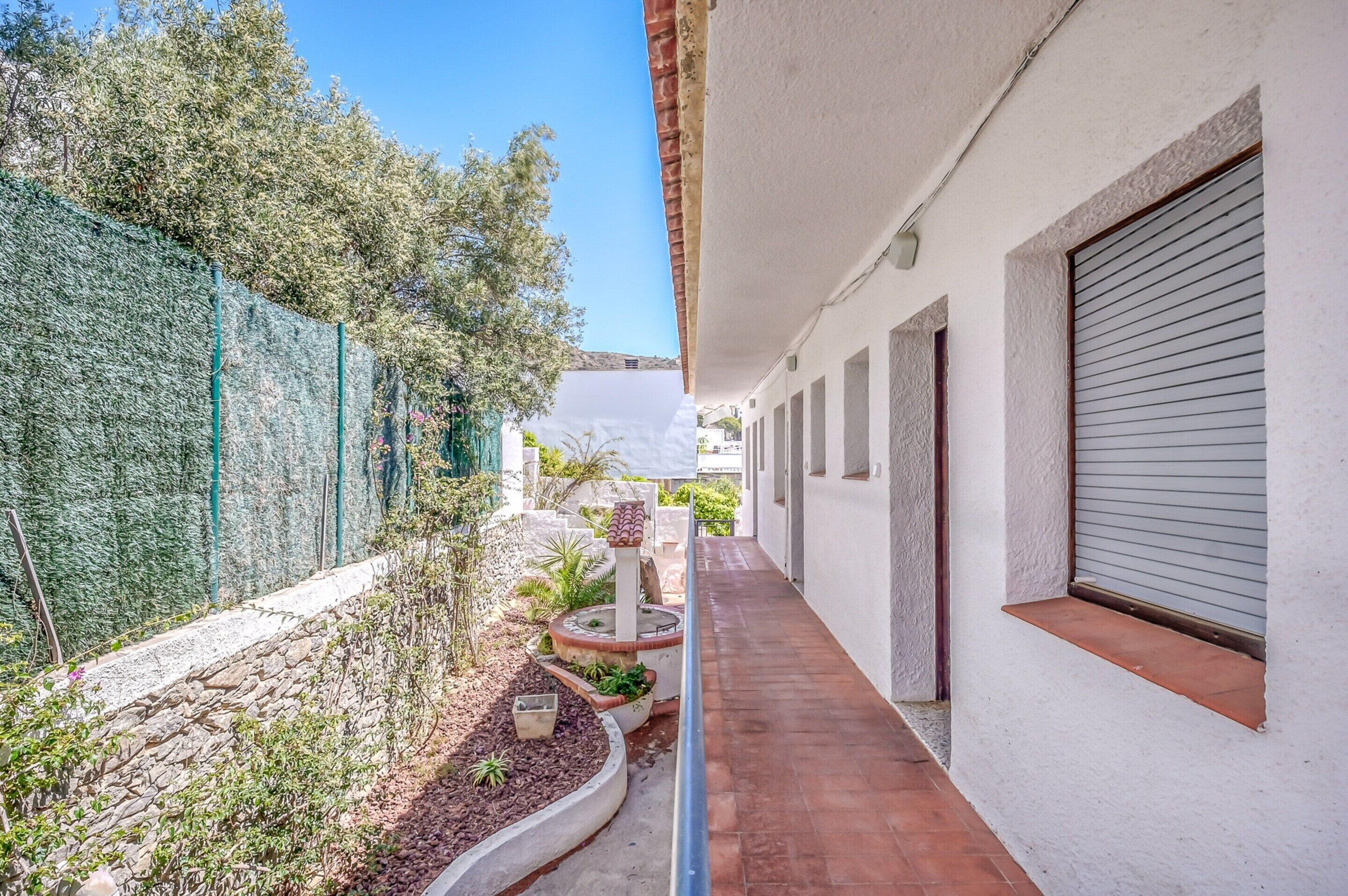 A narrow corridor with a tiled floor, a stone wall on one side, and a white building with a closed shutter on the other.