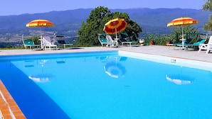 A swimming pool with clear blue water, surrounded by lounge chairs and umbrellas, with a mountainous backdrop. - Farmhouse in Poppi With Pool & Gardens (Poppi)