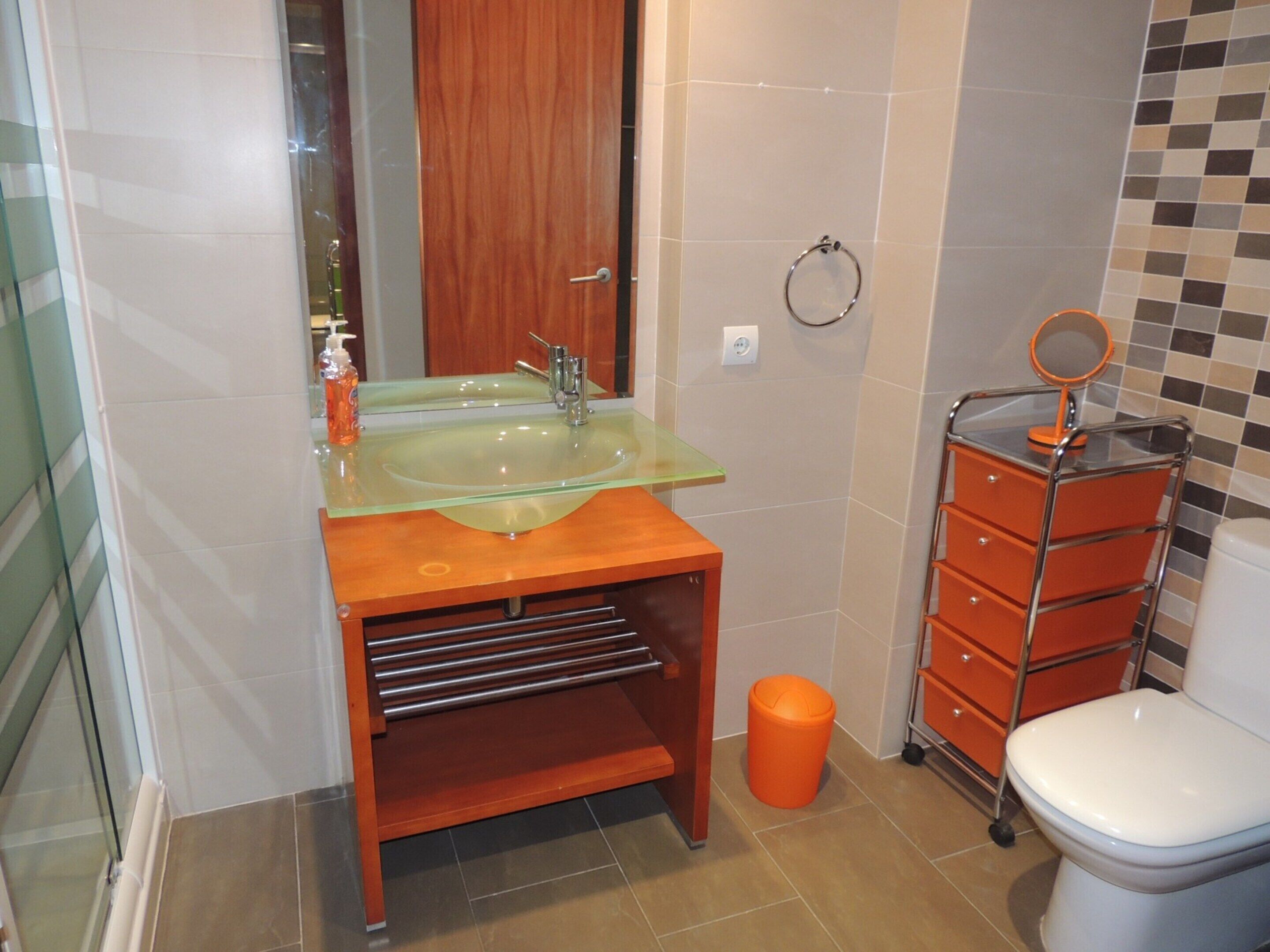 Et badeværelse med vask i glas, træbordplade, orange skraldespand og et hvidt toilet.