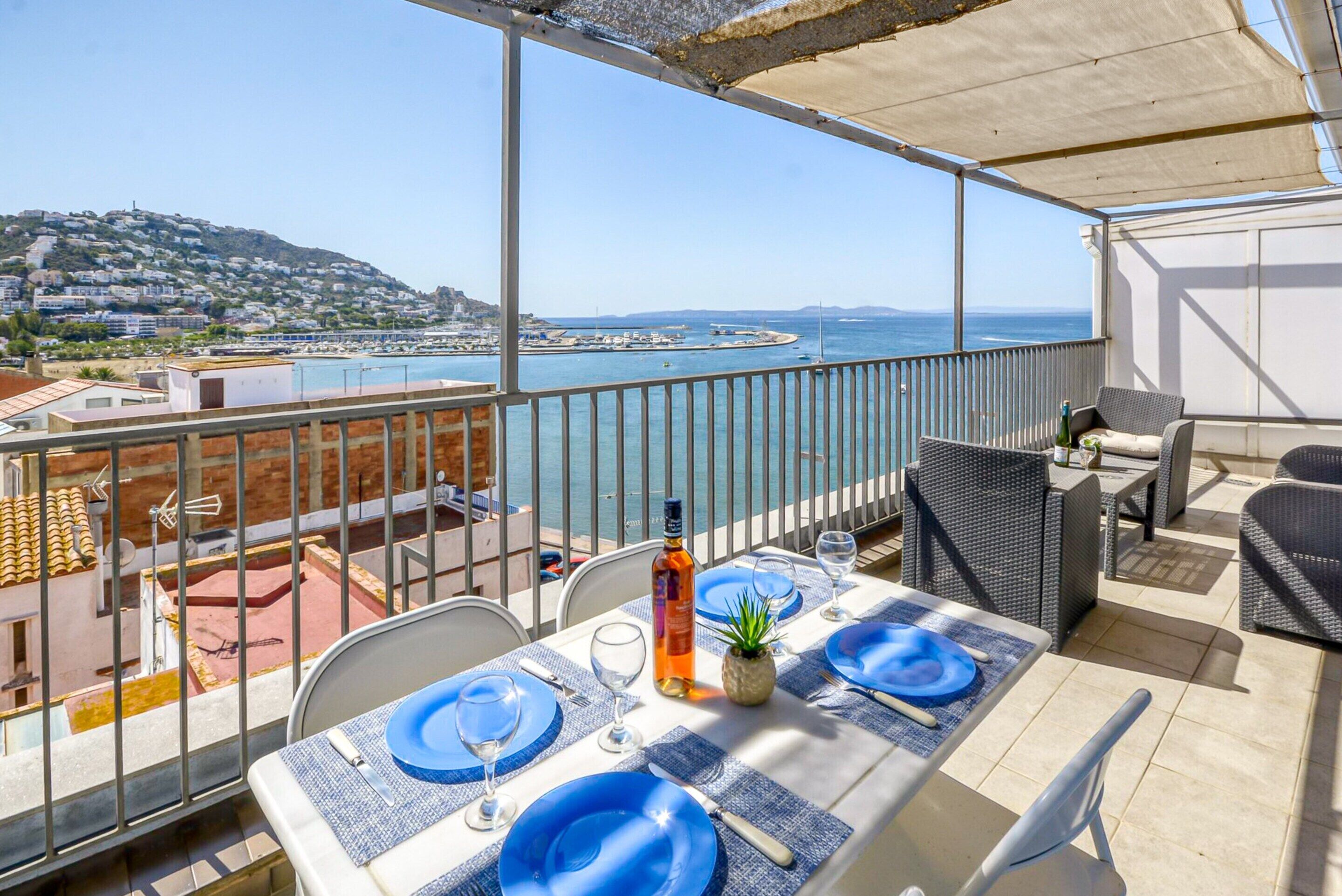 A balcony with a table set for two, overlooking a coastal view with buildings and a beach.