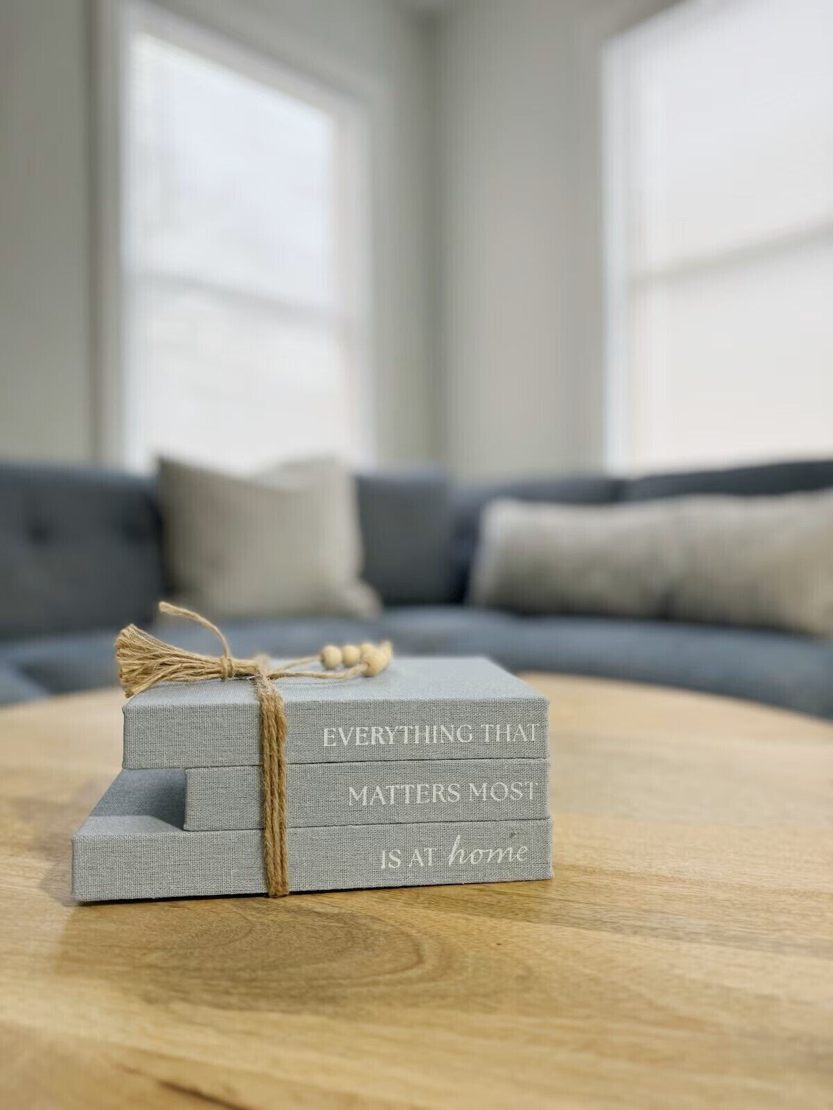 Eine Stapel grauer Stoffkisten mit dem Aufdruck "EVERYTHING THAT MATTERS MOST IS AT HOME" auf einem Holztisch in einem Raum mit grauem Sofa und Fenster im Hintergrund.