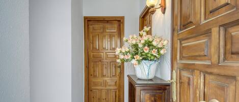 A hallway with a wooden door, a wooden cabinet with a vase of flowers, and a wall-mounted light fixture.