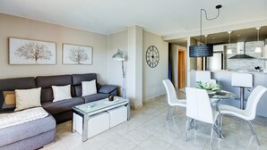 Ein modernes Wohnzimmer mit einem grauen Sofa, einem Glastisch und weißen Stühlen. Es gibt einen Essbereich mit einem weißen Tisch und Stühlen, und im Hintergrund eine Küche.
