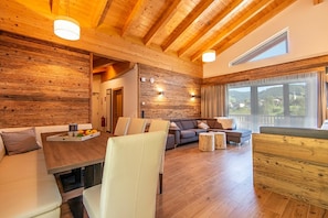 A spacious living area with a wooden ceiling, a sofa set, a dining table with chairs, and a view of the outdoors through large windows.