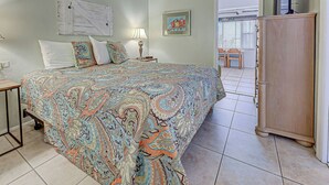 A bedroom with a large bed, a nightstand, a lamp, a wooden cabinet, and a door leading to a balcony. - Directly on the Gulf - Unit  9 - 1 Bedroom - Ground Floor - Sleeps up to 4 (Venice)