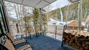 A covered outdoor seating area with chairs and a table, a stack of firewood, and a view of a snowy landscape with trees and buildings. - Private Hot Tub, Views, Summer Adventures (Keystone)