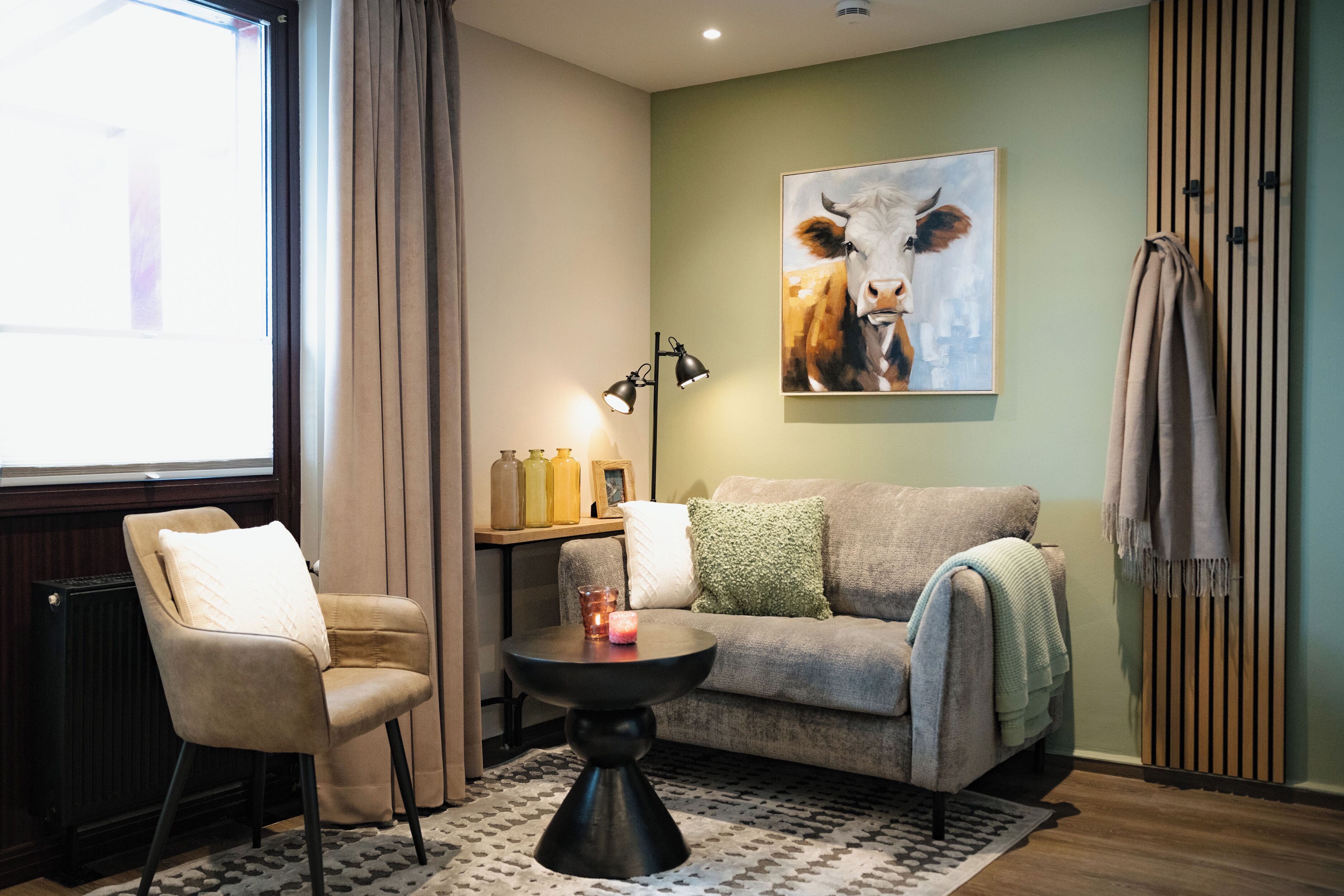 A cozy living room with a beige sofa, a black coffee table, and a chair. A painting of a cow is on the wall, and there's a radiator on the right.