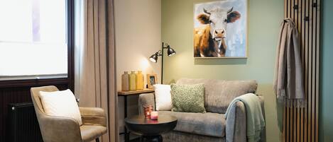 Un salón acogedor con un sofá color beige, una mesa de centro negra y una silla. Un cuadro de una vaca adorna la pared, y hay un radiador a la derecha.