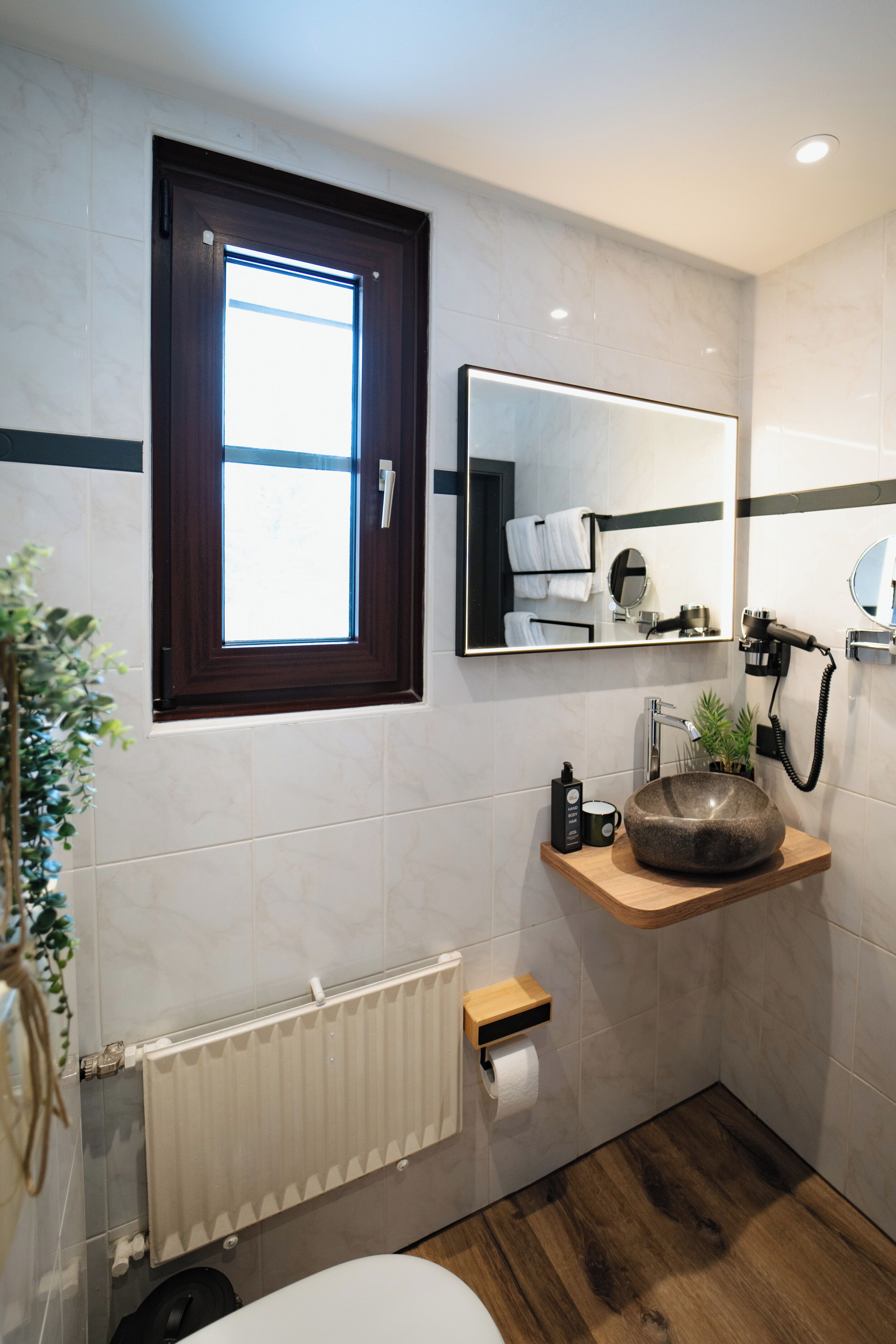 Un baño con tocador de madera, un lavabo redondo, un espejo, un perchero para toallas, un radiador y una ventana.
