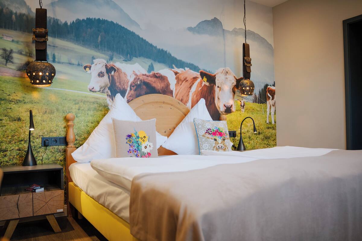 Un dormitorio con cama, almohadas, una mesita de noche y un mural en la pared con vacas en la montaña.