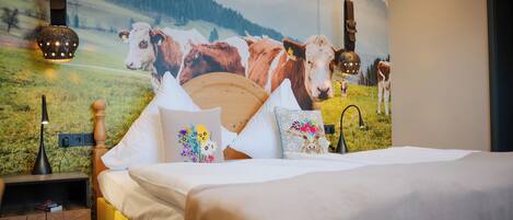 A bedroom with a bed, pillows, a nightstand, and a wall mural of cows on a mountain.
