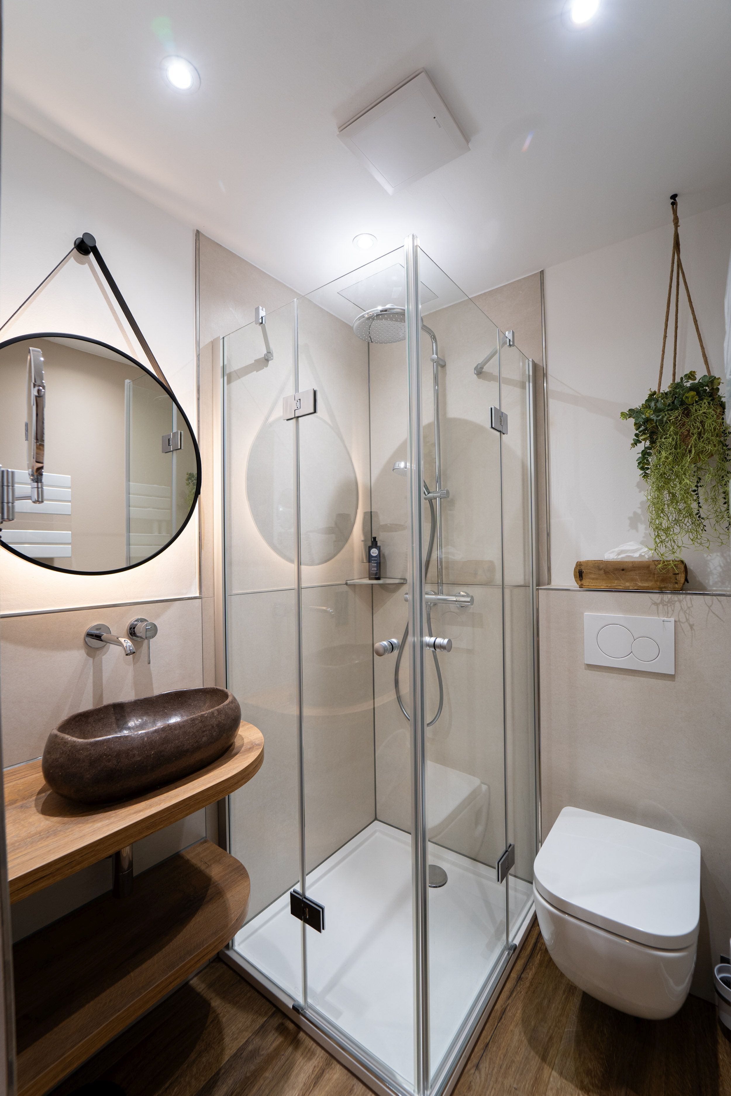 A modern bathroom with a glass-enclosed shower, a wooden vanity, a round mirror, and a white toilet.