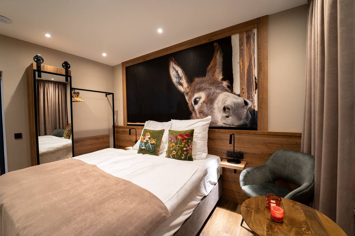 Una habitación de hotel con una cama grande, cabecera de madera, una obra de arte colgada de un burro, un escritorio con una silla y una pequeña mesa redonda con una vela.