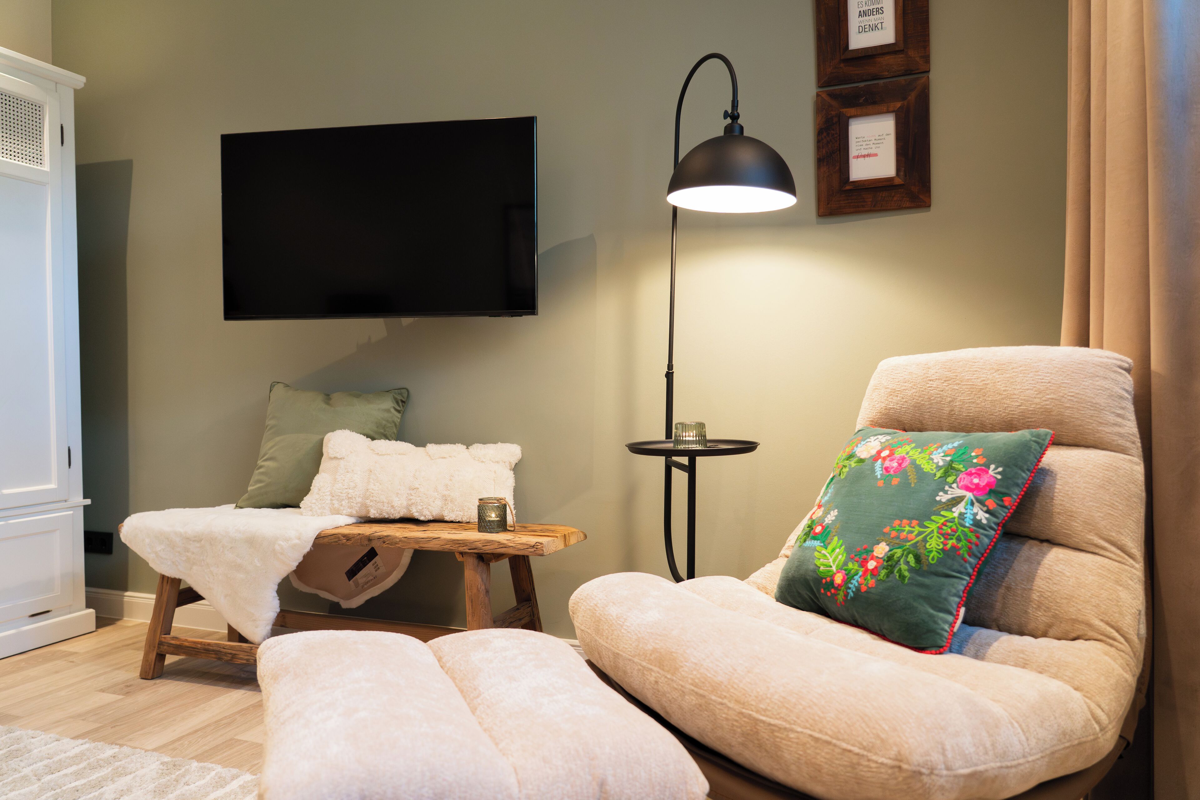 Ein gemütliches Wohnzimmer mit einem Flachbildfernseher, einem bequemen Sessel mit floralem Kissen, einem kleinen Holztisch mit Decke und Kerze sowie einer Stehlampe.