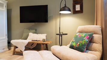 Ein gemütliches Wohnzimmer mit einem Flachbildfernseher, einem bequemen Sessel mit floralem Kissen, einem kleinen Holztisch mit Decke und Kerze sowie einer Stehlampe.