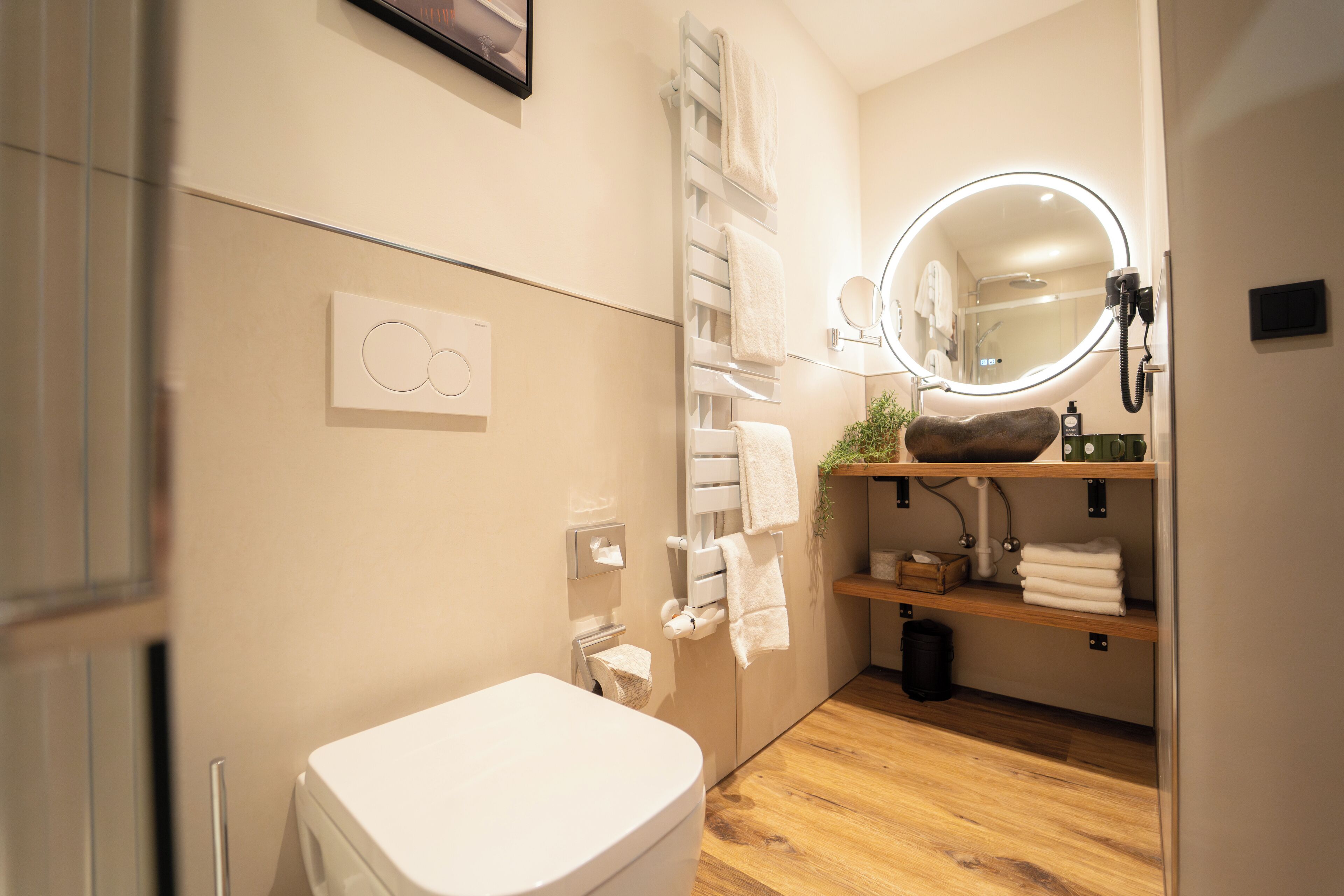 A modern bathroom with a toilet, a towel rack, a mirror with a ring light, and a shelf with towels.