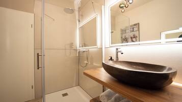 Un baño moderno con ducha envuelta en vidrio, un tocador de madera con un lavabo tipo cuenco grande e iluminación integrada en el espejo.