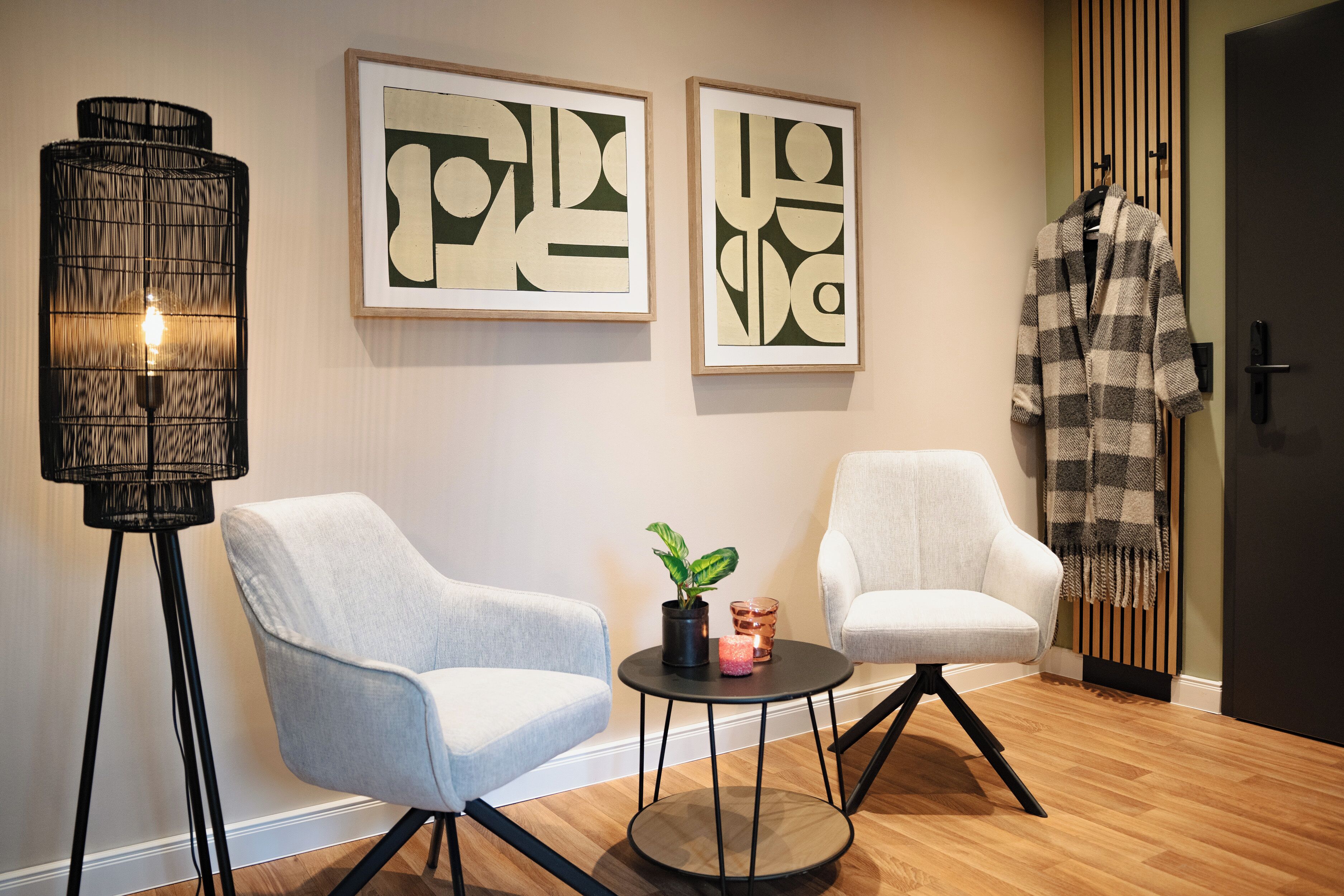 A room with two armchairs, a small round table with a plant and candles, a tall floor lamp, and two framed abstract artworks on the wall.
