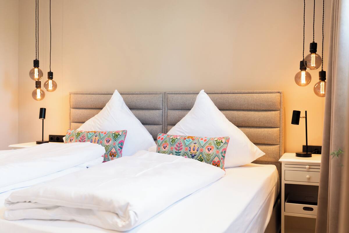 Una cama bien tendida con sábanas blancas, dos almohadas decorativas, una mesita de noche con una lámpara y bombillas colgantes.