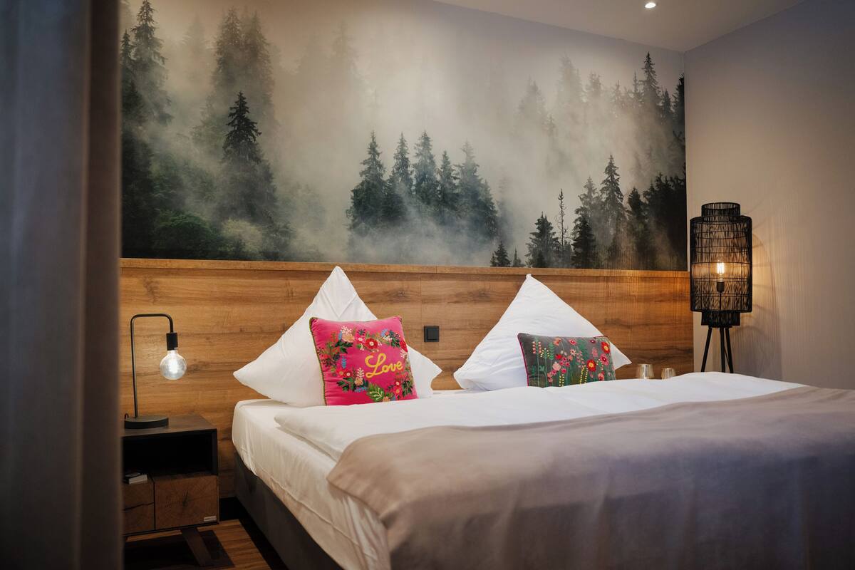 Un dormitorio con cabecera de madera, cama con ropa de cama blanca, dos almohadas decorativas, una mesita de noche con lámpara y un mural en la pared con un bosque.