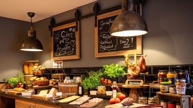 A kitchen counter with various food items, including meats, vegetables, and condiments. There are chalkboards with text, hanging lights, and wooden cabinets.