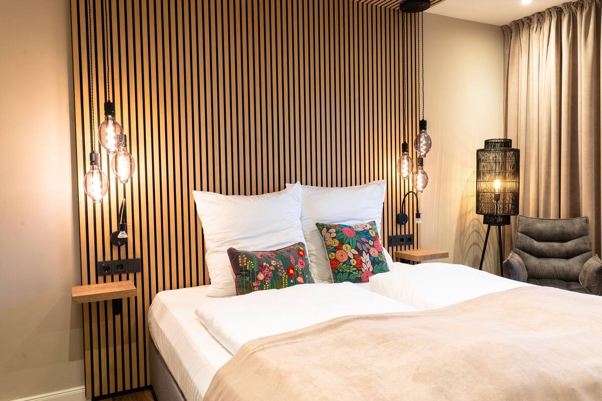 Una cama bien hecha con una colcha color beige, dos almohadas con motivos florales y un cabecero de listones de madera. Hay lámparas de noche con bombillas a la vista y una lámpara de pie con pantalla tejida. Un sillón gris está ubicado a la derecha de la cama.
