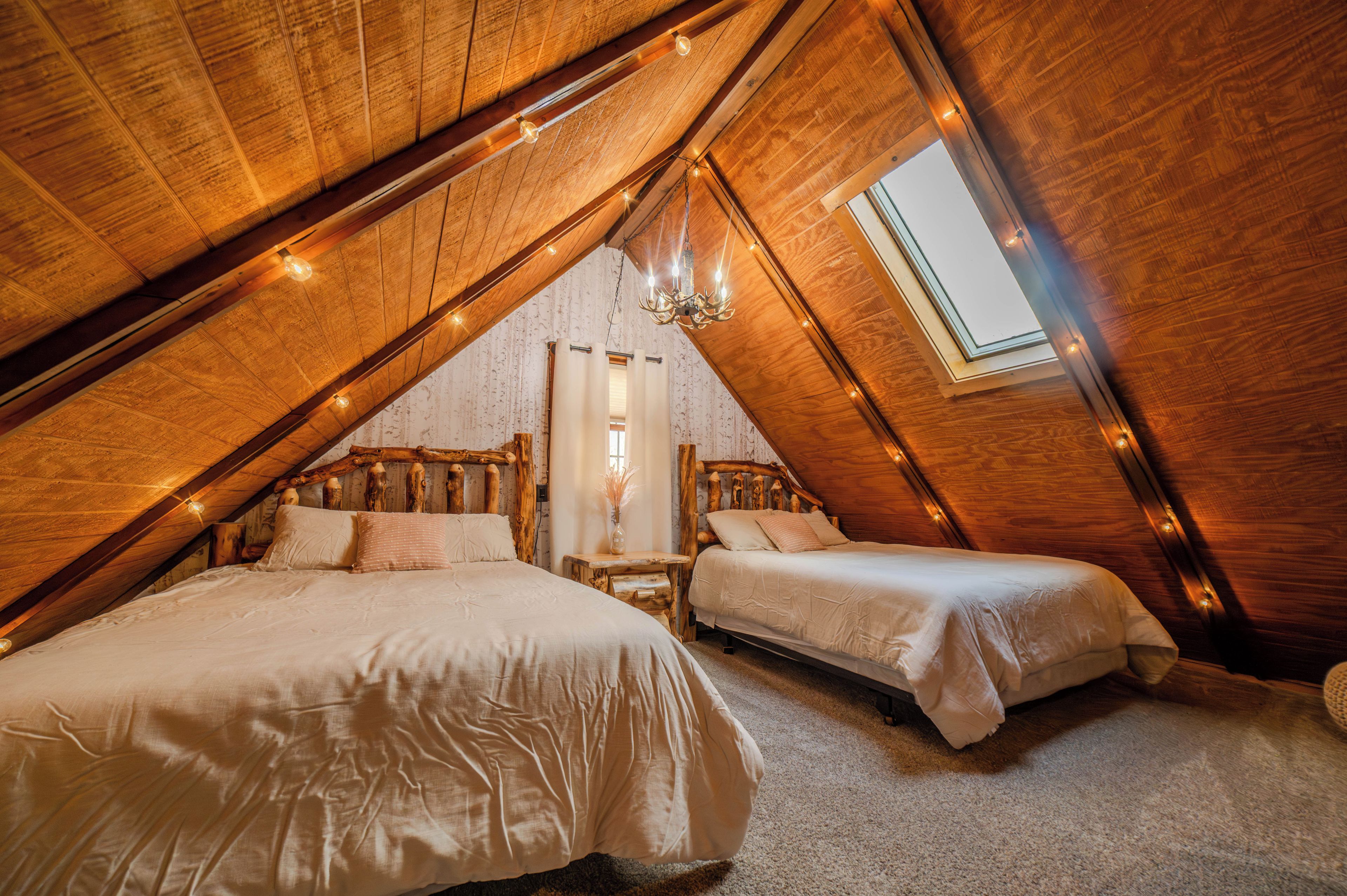 Une chambre mansardée avec deux lits, des poutres en bois, un puits de lumière et des guirlandes lumineuses.
