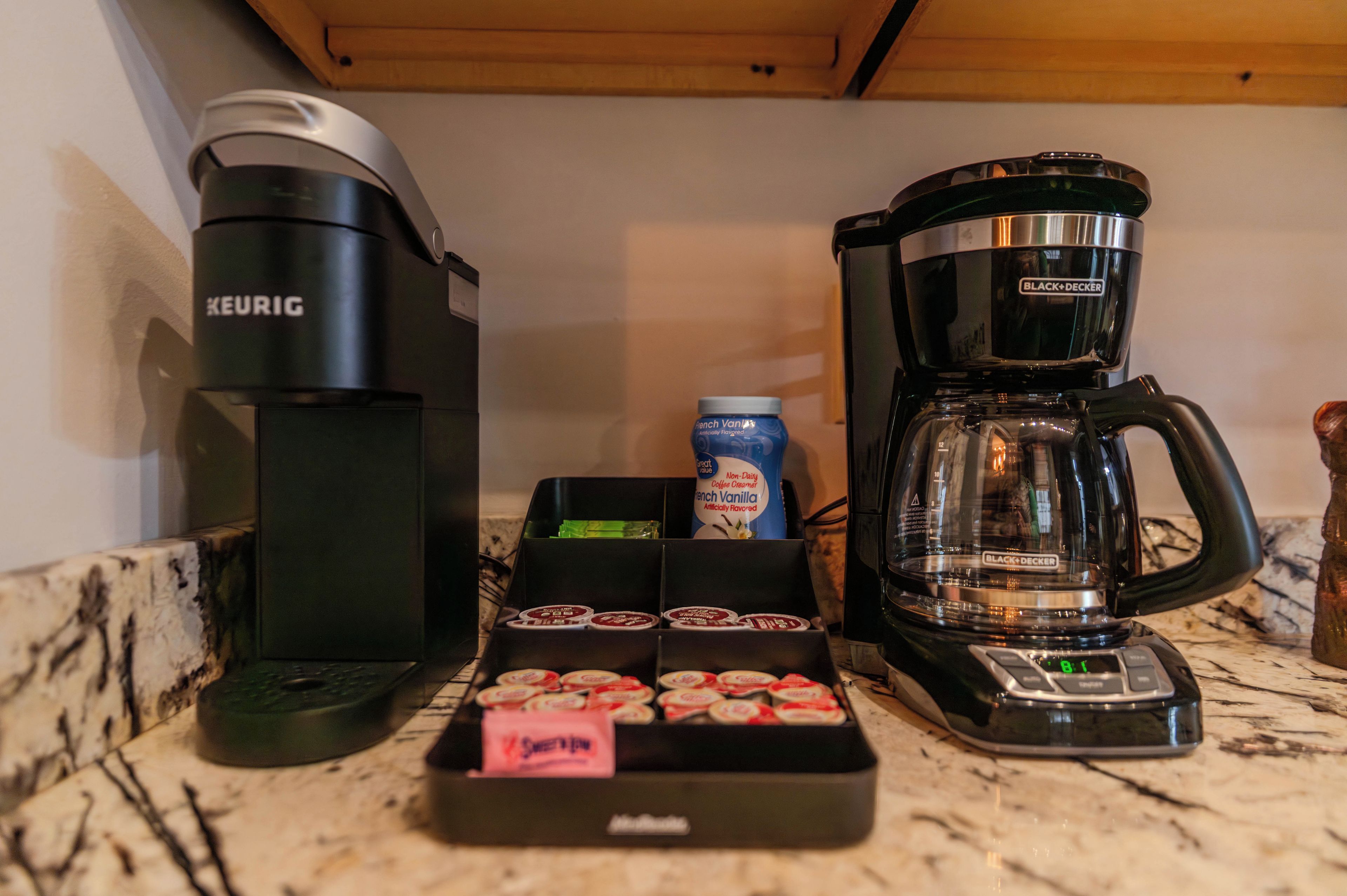 A kitchen with a Keurig coffee maker, a Black+Decker coffee maker, a jar of French Vanilla creamer, and a box of Sweet'n Low.