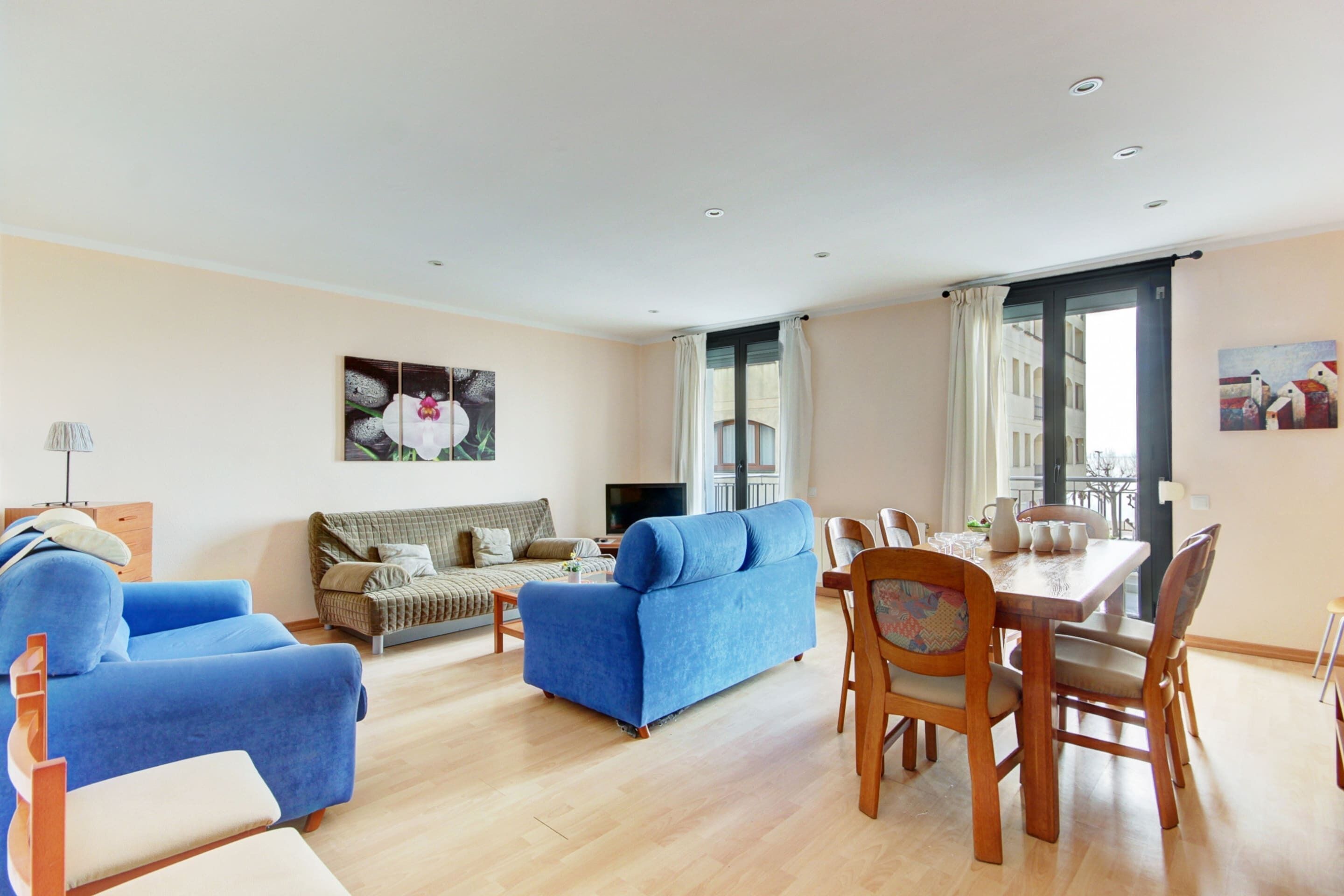 A spacious living room with a blue sofa, a wooden dining table, and a large window.