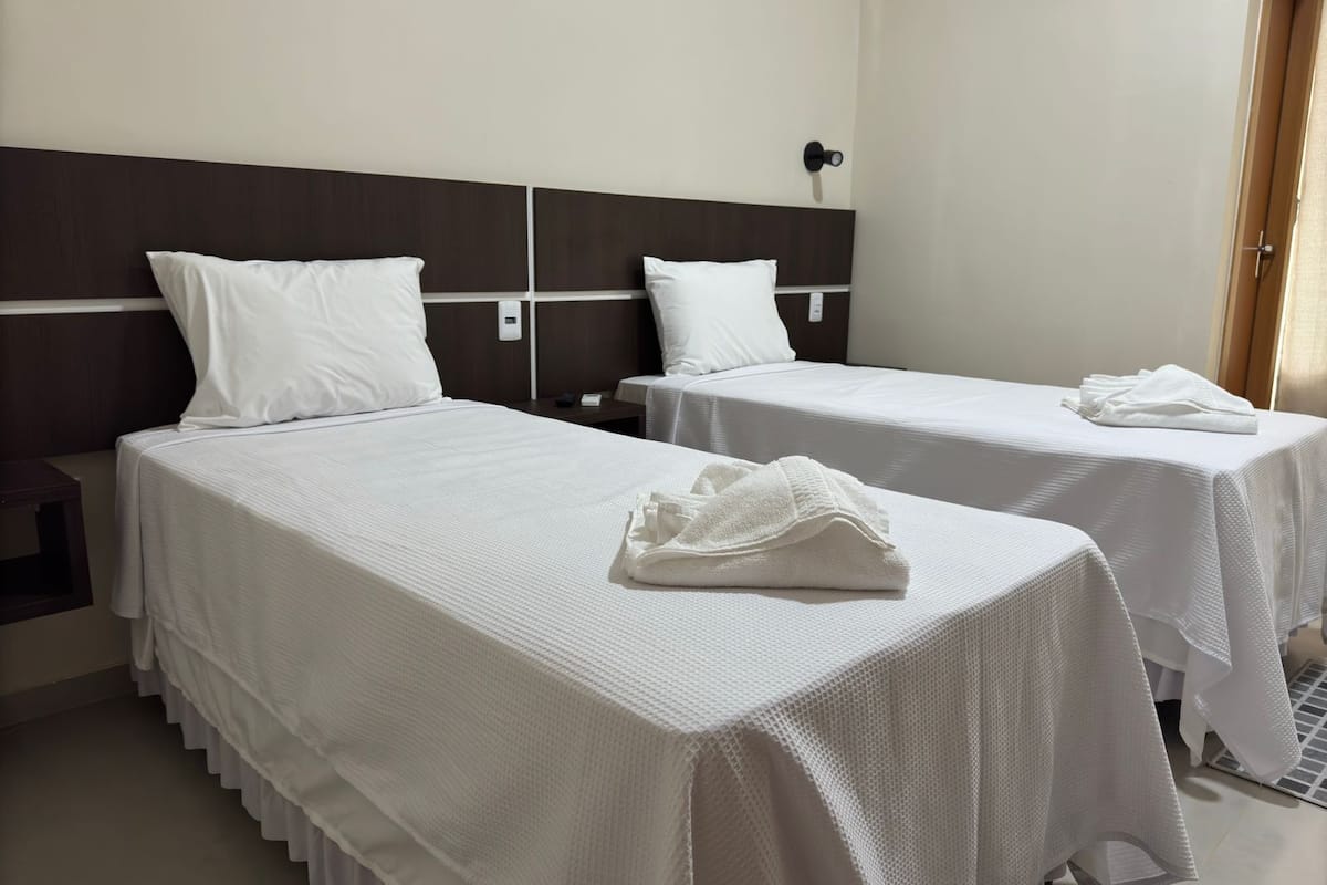 Une chambre d’hôtel avec deux lits simples, chacun recouvert d’une courtepointe blanche et agrémenté d’oreillers, une tête de lit en bois foncé, une table de chevet avec un téléphone et une serviette pliée posée sur le lit.