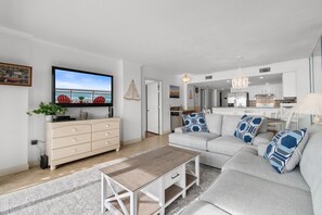 Ein modernes Wohnzimmer mit Sofa, Couchtisch und Fernseher. Im Hintergrund ist eine Küche zu sehen.