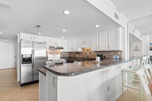 Eine moderne Küche mit weißen Schränken, einem Edelstahl-Kühlschrank, einer Kücheninsel mit Granitplatte und einem Essbereich mit weißen Stühlen.