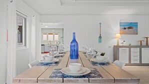 A dining area with a wooden table set for four, a blue vase centerpiece, and a kitchen in the background. - 4-Min to Eagle Beach* Private Pool* (Noord)