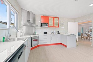 A modern kitchen with white cabinetry, a red accent wall, stainless steel appliances, and a view of the living area. - Backyard Bliss: 4BR Pool Home, 4Min to Eagle Beach (Noord)