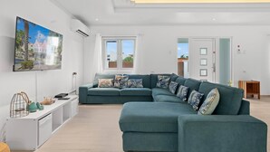 A modern living room with a teal sectional sofa, a flat-screen TV mounted on the wall, and a minimalist design. - 4-Min to Eagle Beach* Private Pool* (Noord)