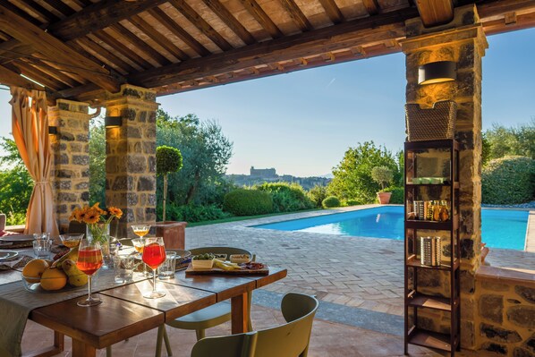 An outdoor dining area with a pool view, stone pillars, and a shelf with bottles. - Villa San Gabriele: Historic Charm and Modern Comfort (Orvieto)
