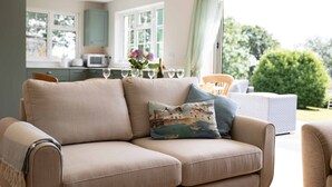 Un canapé beige agrémenté d’un coussin à motifs, une table à manger avec des chaises, un coin cuisine et une vue sur la verdure à l’extérieur.