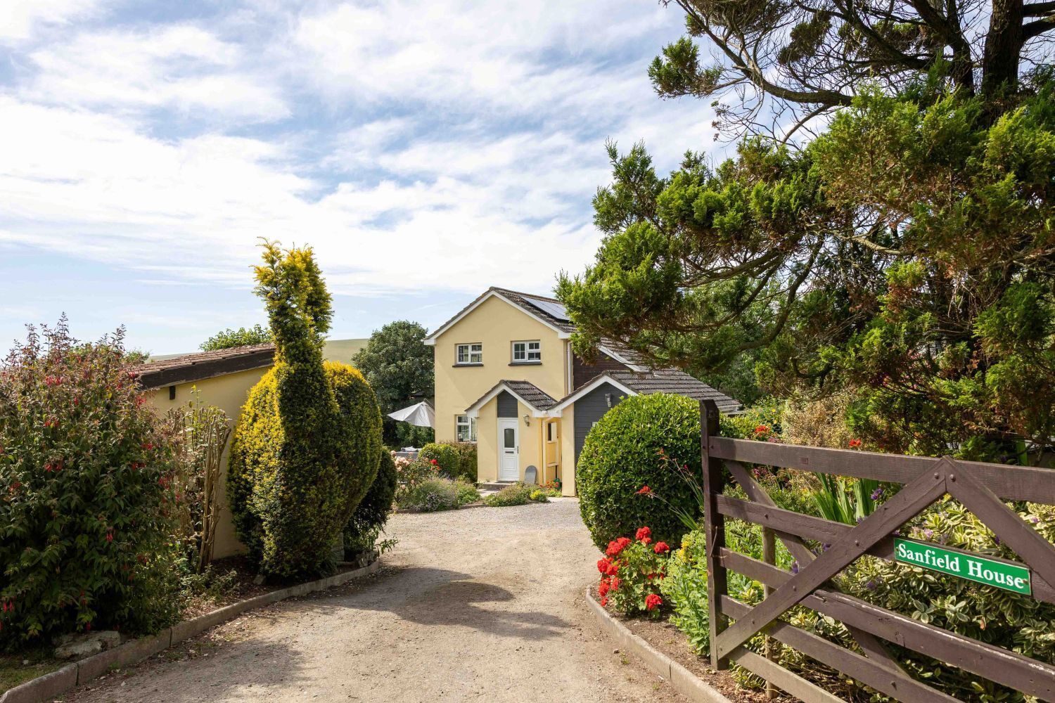 Uma casa de dois andares com uma entrada de cascalho que leva a um portão de madeira com a placa 'Sanfield House'.