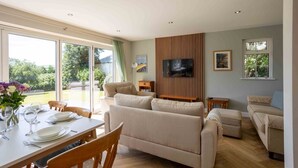 Un vaste espace de vie comprenant un canapé, un téléviseur fixé sur un panneau en bois, une table à manger pour deux personnes et une grande porte-fenêtre coulissante donnant sur un jardin.