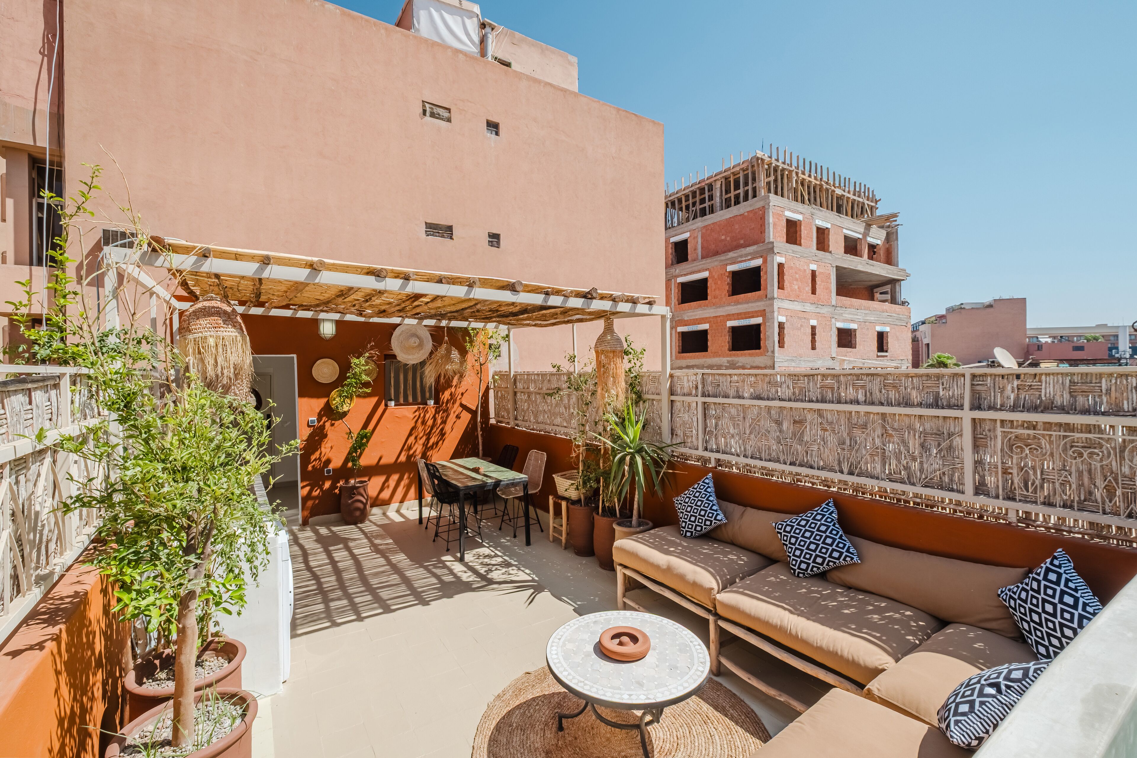 A rooftop patio with a sofa, table, and chairs, surrounded by potted plants and a decorative wall.
