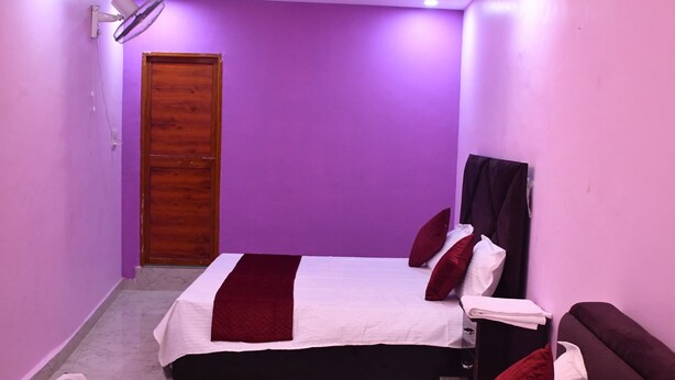 A small, dimly lit room with purple walls, two beds with red and white bedding, a ceiling fan, and a wooden door. - Dev In & homestay (Faizabad)