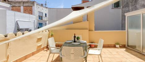 A rooftop terrace with a table set for two, surrounded by white chairs, and a small potted plant.