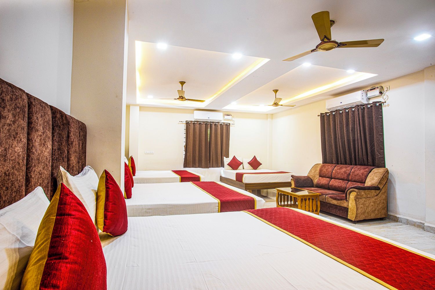A hotel room with two beds, a sofa, a dining table, and ceiling fans.