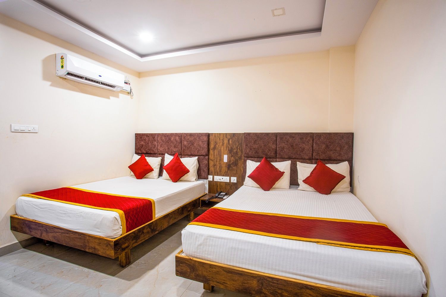 Habitación de hotel con dos camas, cada una con cabecero de madera y ropa de cama roja y blanca. Un equipo de aire acondicionado está montado en la pared sobre las camas.